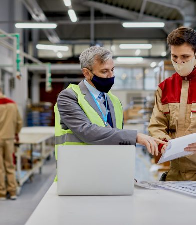 Factory worker and businessman with protective face masks analyzing reports at woodworking production facility.