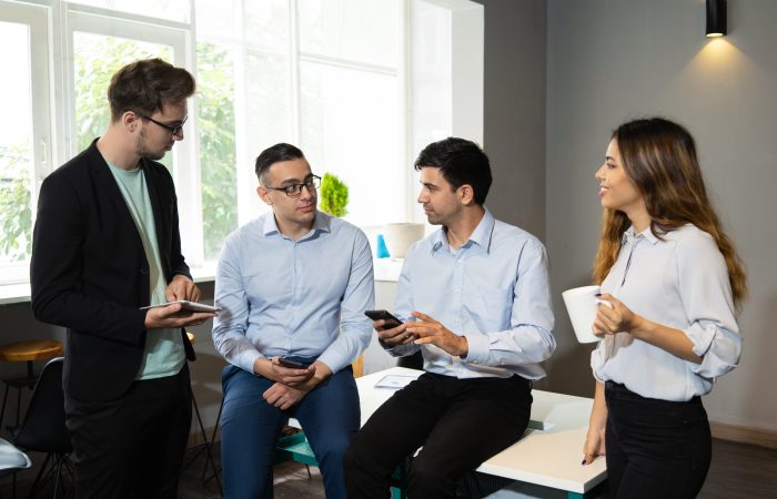 Professional team consulting on internet while discussing ongoing project. Four employees talking in meeting workspace, using gadgets and drinking coffee. Interaction concept